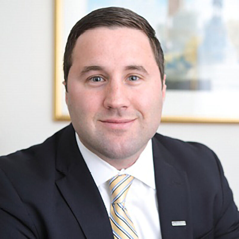 The image shows a man in a suit and tie. He has short brown hair and is looking at the camera with a slight smile. The background is blurred, but it appears to be an office setting with a painting on the wall. The man looks to be in his late 20s or early 30s.